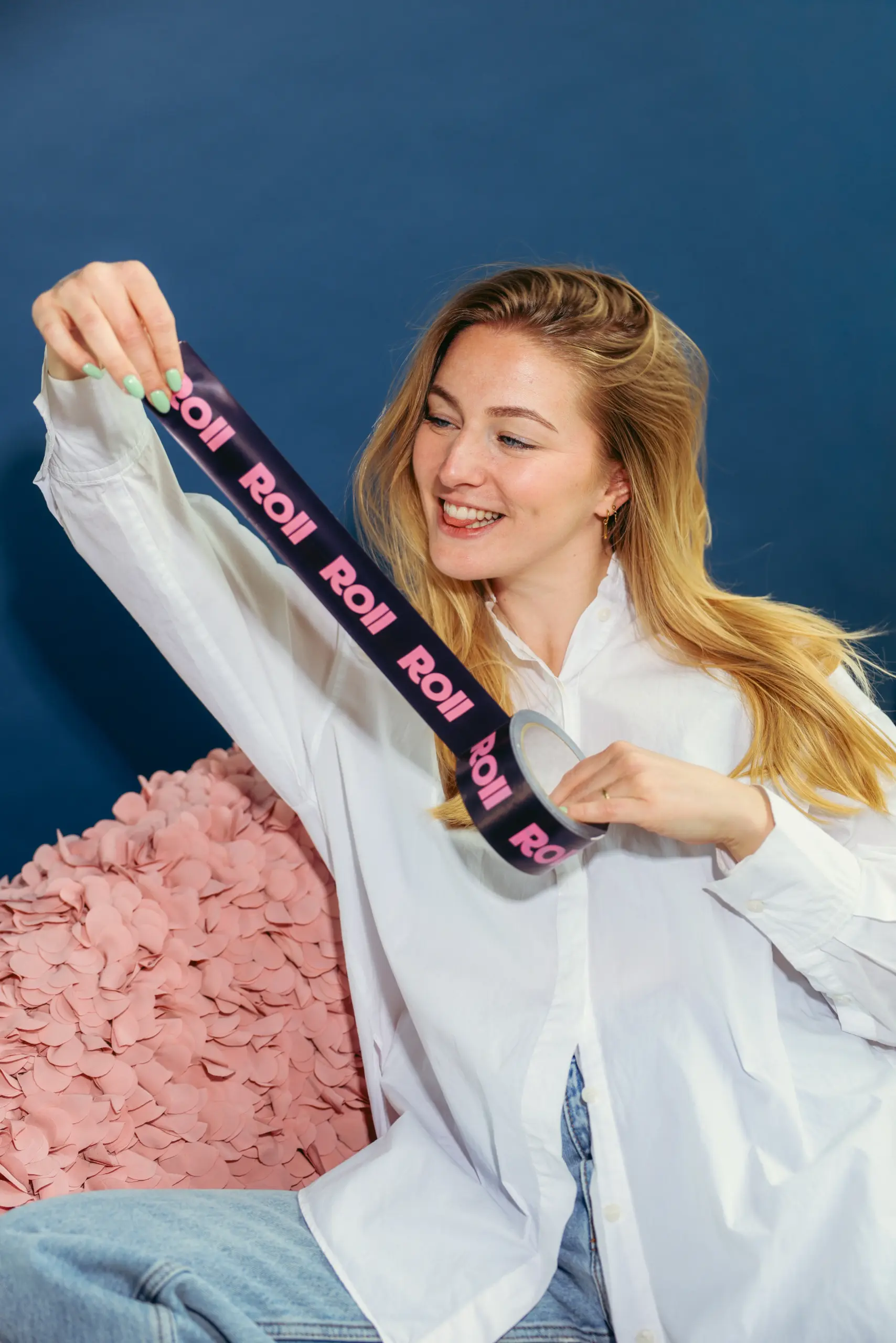 Vrouw trekt lachend een rol donkerpaars plakband af met roze Roll-logo's, zittend tegen een blauwe achtergrond met een roze sierkussen.
