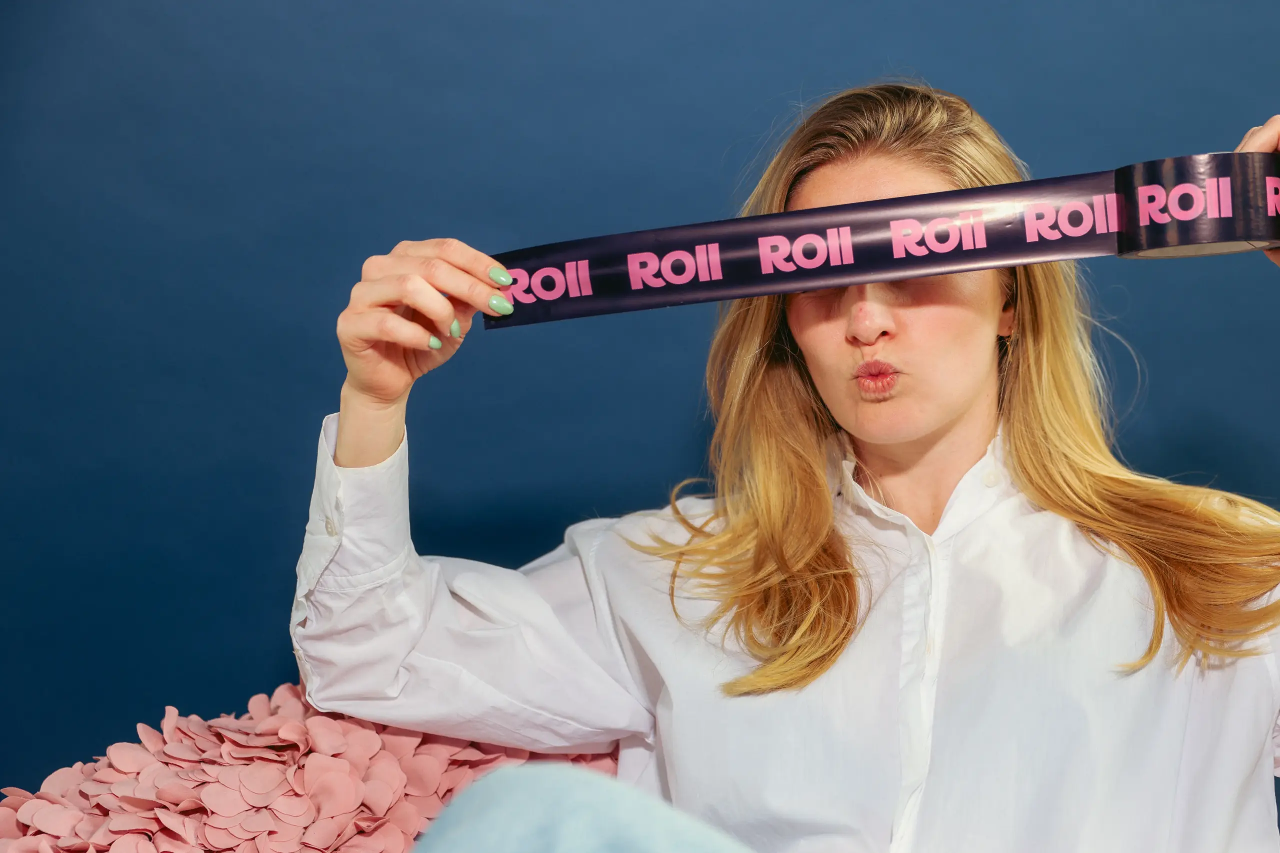 Vrouw met blond haar houdt een donkere Roll-tape voor haar ogen terwijl ze een kusmond maakt, omgeven door roze confetti.